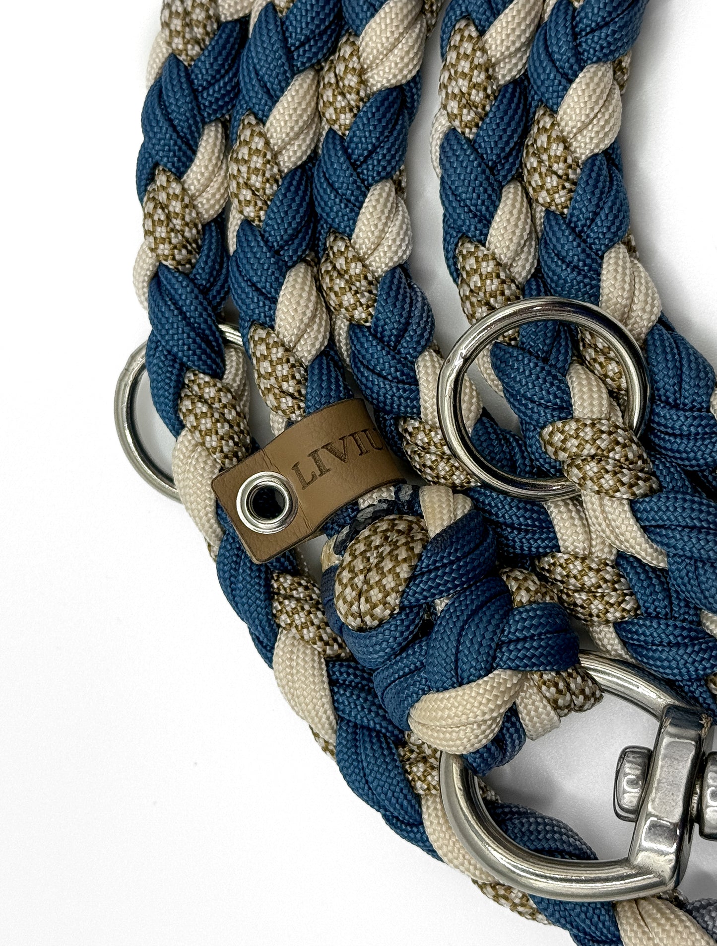 Handgefertigte Hundeleine aus robustem Paracord in Blau, Beige und Naturtönen mit 2 stabilen Metallkarabinern und 3 Metallringen – hochwertig und langlebig. Es sind 2 Ringe zu sehen, ein Logolabel und ein Teil des Karabiners.
